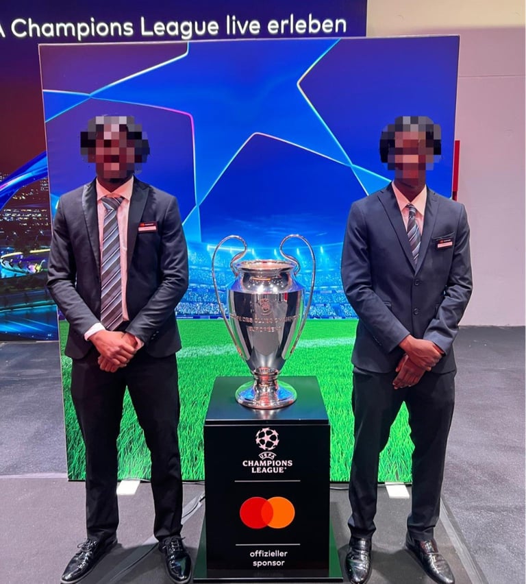 Two men in business suits stand beside the UEFA Champions League trophy at a live event display with a stadium background